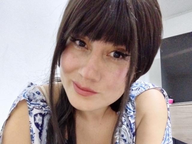 AngelCams has brown hair with bangs, wearing a blue and white patterned top, showing a slight smile with pink lips, and has a cute, feminine face.