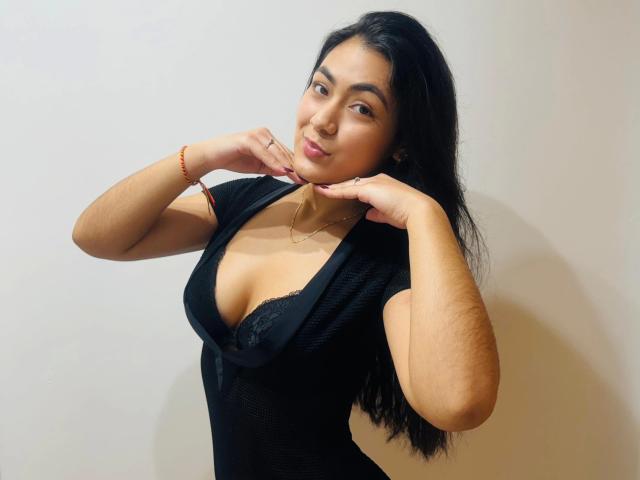 JenniferMet-hot with dark hair, medium tits, wearing a black, deep V-neck top, hands near face, light skin, slight smile, red bracelet, necklace, and subtle makeup.