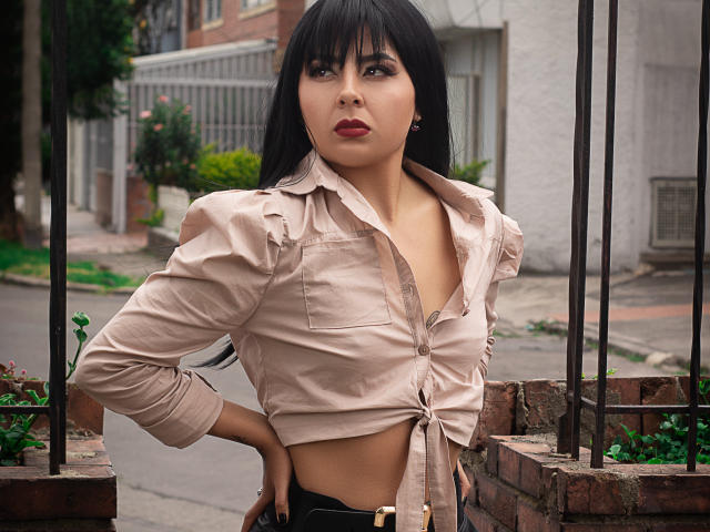 NebulosaI with black hair, tied beige shirt, exposed midriff, hands on hips, looking up with parted lips, standing in front of a brick fence, wearing black pants.