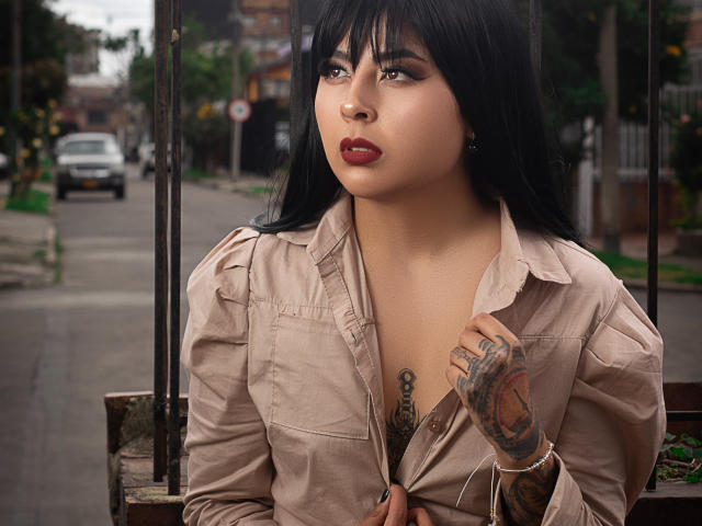 NebulosaI with black hair, red lipstick, and tattoos on her chest and hand, wearing an unbuttoned beige shirt, sits on a bench, holding her shirt.