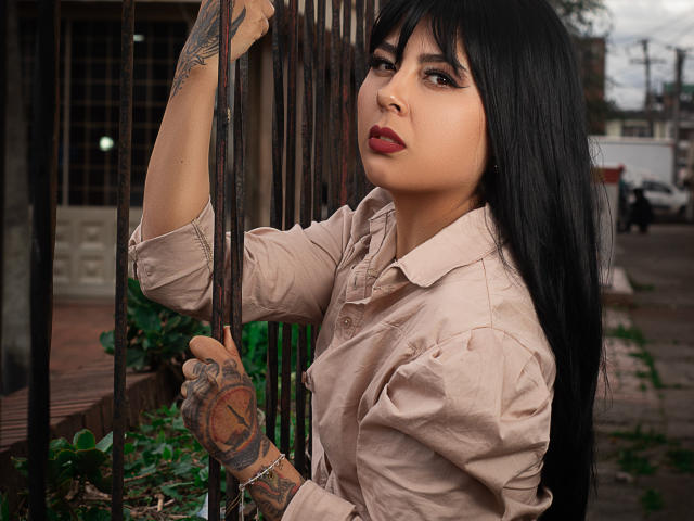 NebulosaI with long black hair, tan button-up shirt, and tattoos on her right hand and left forearm, holding a black metal fence, looking over her shoulder with red lipstick.