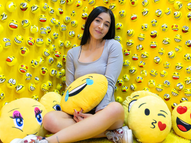 NicoColeman, with black hair and light skin, sits cross-legged, holding a yellow emoji pillow, surrounded by yellow emoji wall decorations, wearing a gray long-sleeve top and white sneakers.