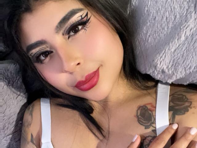 NebulosaI with dark hair, thick eyebrows, red lipstick, and white tank top, showing tattoos on her chest, lying on a grey pillow, looking at the camera.