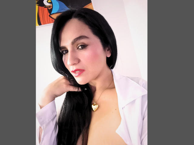 CameronViviane with black hair, pink cheeks, red lipstick, white shirt open, showing cleavage, gold necklace, holding hair, looking at camera, grey background, cartoon partially visible.