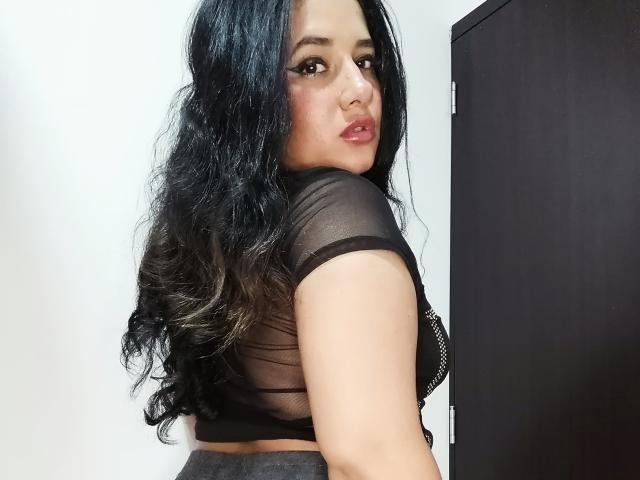 Ansol with long black wavy hair, wearing a sheer black top, showing her ass and looking over her shoulder with a slightly open mouth. She's standing by a dark door.