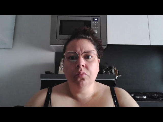Lamante a un visage rond, porte des lunettes et une nuisette noire à bretelles fines, avec ses seins légèrement visibles, dans une cuisine où l'on voit un micro-ondes au-dessus..