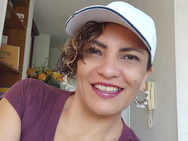 ShayFoxxy with curly brown hair, white cap, purple shirt, pink lipstick, smiling, medium tits, earring with coiled wire, standing in a room with yellow flowers.