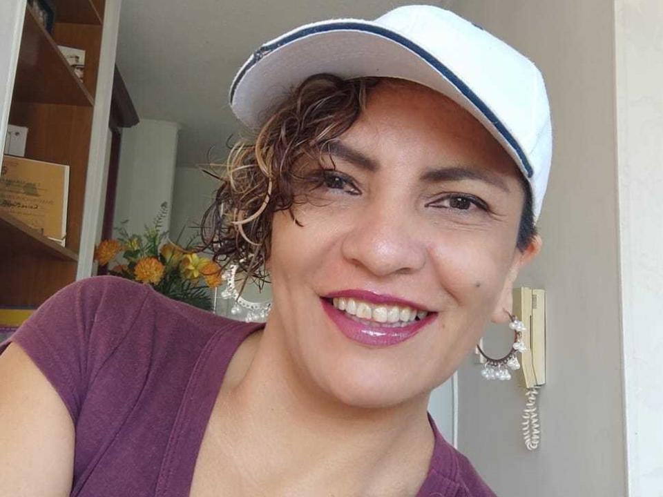 ShayFoxxy with curly brown hair, white cap, purple shirt, pink lipstick, smiling, medium tits, earring with coiled wire, standing in a room with yellow flowers.