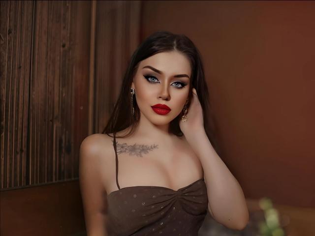 MissNestie with blue eyes, red lipstick, dark brown hair, wearing a brown, strapless, polka-dotted top, showing her tits and a tattoo above her chest. Her hand touches her hair.