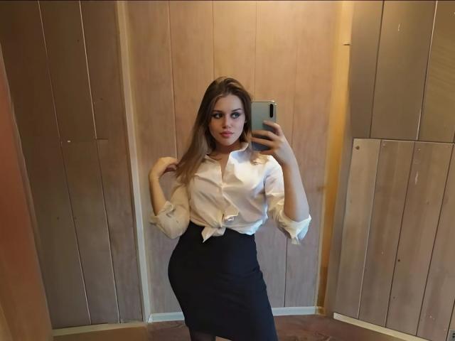MissNestie with long brown hair, wearing a tied white shirt and black skirt, holding a phone, standing in front of wooden panels, showing her ass in a mirror selfie.
