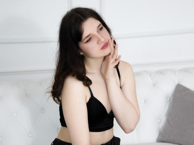 BonnieFlower with pale skin and dark brown hair, wearing a black bra, touching her cheek, eyes closed, sitting on a white tufted couch, looking sexy and relaxed.
