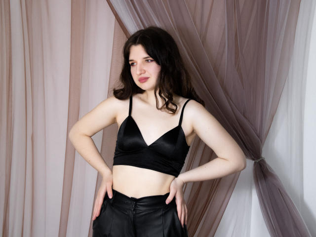 BonnieFlower with pale skin and dark wavy hair, wearing a black bra and black leather pants, standing with hands on hips, looking slightly to the side.