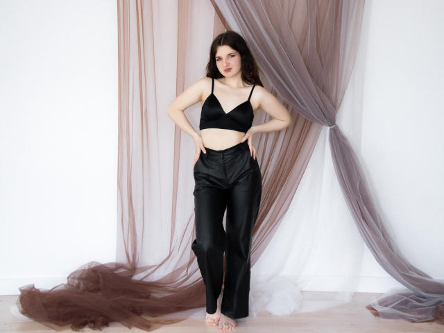BonnieFlower stands barefoot with hands on hips, wearing a black bra and shiny black pants. She has long brown hair and a slight smile. Her tits are small, pussy is hidden.