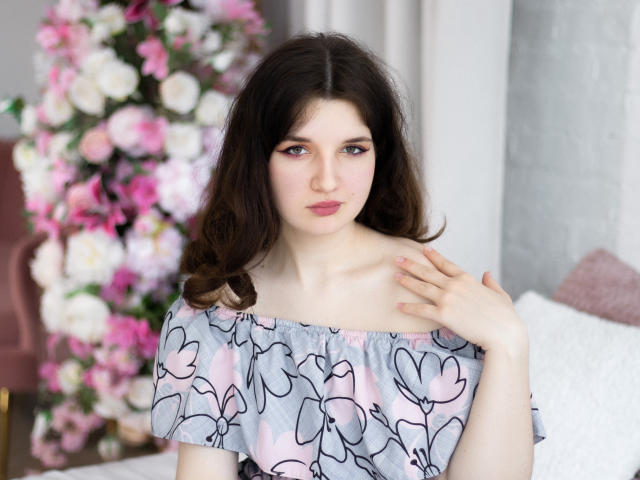 BonnieFlower has pale skin, dark wavy hair, and pink lips. She wears a floral off-shoulder top, hand on collarbone, with pink and white flowers in the background.