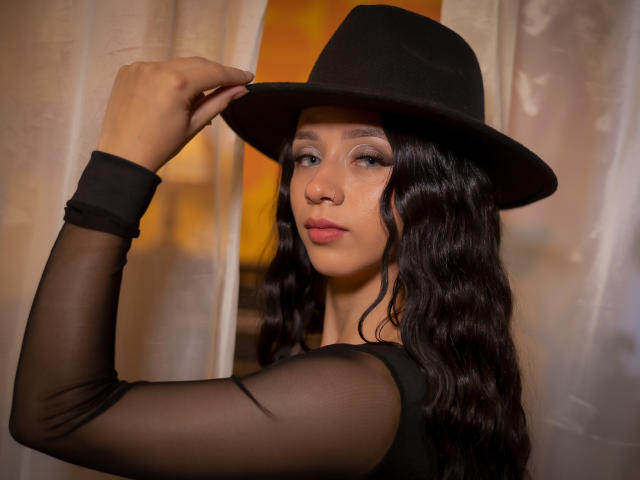 KloeThompson with wavy black hair, wearing a black sheer long-sleeve top and black hat, touching the brim, looking sideways, soft yellow light in background.