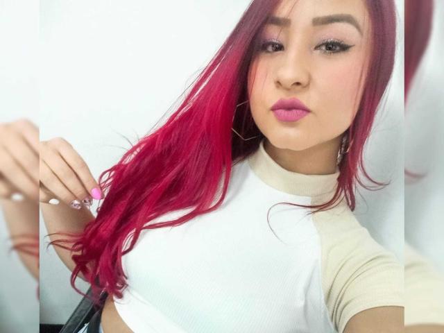 MagicAmberVincent with bright pink hair, white rag shirt, pink lipstick, and white nails, holding her hair. Her boobs are small, and her ass is not visible.