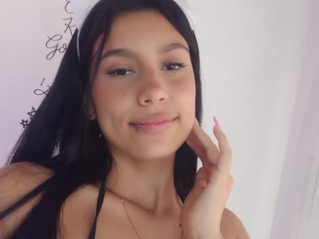 KataleyaMansson has long black hair, brown eyes, and a slight smile with a tongue piercing. She's touching her cheek with pink nails, wearing a black halter top.