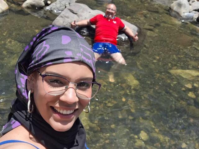 AlexKaren with glasses and a purple headscarf smiling, wearing a blue top, foreground. Bald man in red shirt, blue shorts, sitting in clear water, background.