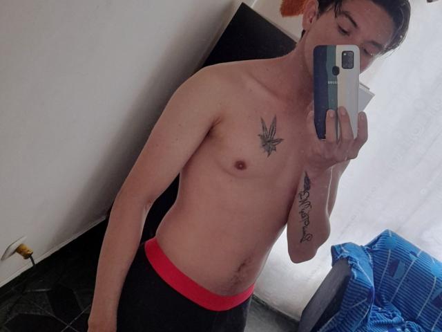 FlacoVergon69 is shirtless with a small tattoo of a marijuana leaf on his chest and a cursive tattoo on his right forearm. He's wearing black and red underwear.