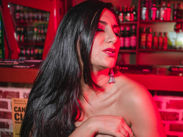 JuliaRobinson with long black hair, red-lit skin, and pink lipstick, covering her bare tits with one arm, wearing a silver chandelier earring, eyes closed in a bar.