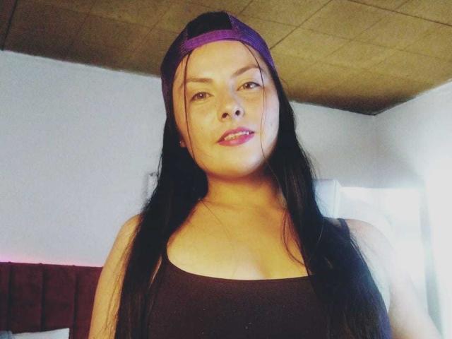 BrandySweet, Asian woman with long black hair, purple backwards cap, and black tank top, showing her medium-sized tits, smiling slightly, in a dimly lit room.