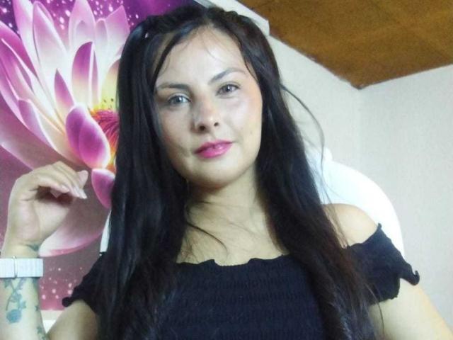 BrandySweet with long black hair, pink lipstick, and black off-shoulder top, holding a pink flower, wearing a white wristwatch, tattoo on left wrist.