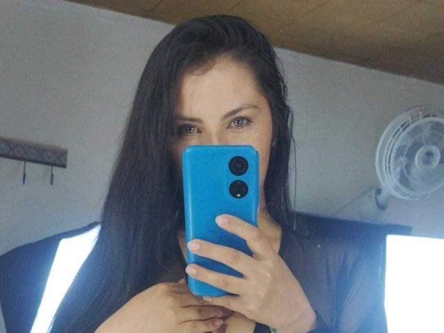 BrandySweet with long black hair, holding a blue phone, covering her tits, mirror selfie, wooden ceiling, white fan, bright lights, looking at the camera, focused expression.