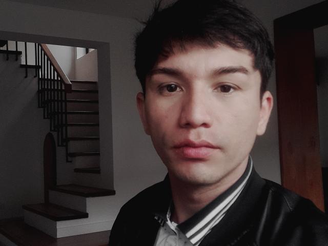 FlacoVergon69, with short dark hair and serious expression, wearing a black leather jacket over a striped shirt, stands in front of a wooden staircase.