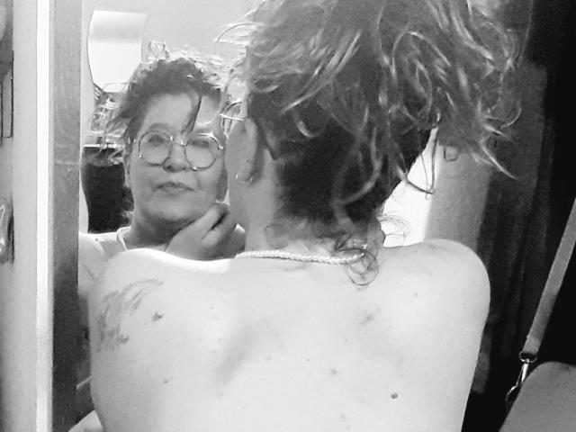 Choukette com cabelo cacheado e molhado, de costas para o espelho, mostrando a parte superior das costas e uma pequena tatuagem, rosto refletido com óculos, sorriso leve, mão perto do queixo, foto em preto e branco..