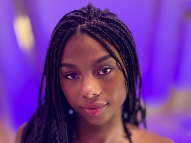 EbonyMore heeft een donkerbruine huid, een kleine neus en lang zwart gevlochten haar. Ze glimlacht met roze lippen, draagt een neusring en heeft een paars, onscherpe achtergrond..