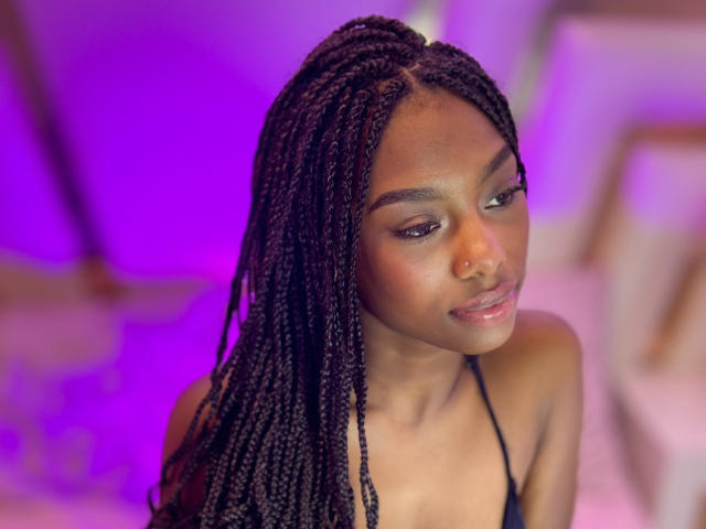 Ebony, met donkere huid, lange zwarte vlechten en een kleine neusring, kijkt naar beneden met een subtiele glimlach. Ze draagt een zwarte haltertop, en er is paars licht..
