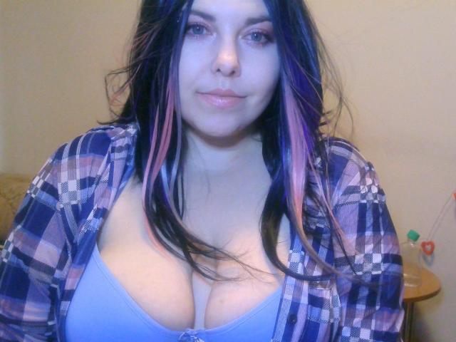 WhitteLotus with long black and pink-streaked hair, wearing a blue bra and checkered shirt, showing her big, round, ample tits. She has a subtle, cute smile.