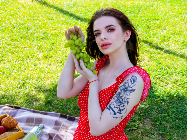 AdeleClifford with green grapes, wearing a red polka dot dress, holding them up, showing a black floral tattoo on her right arm, sitting on a checkered blanket.