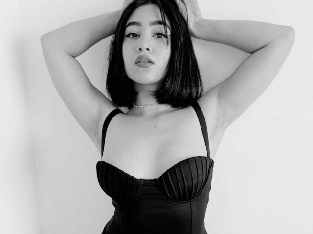 Annakomatsu, mit erhobenen Armen, trägt ein schwarzes, gerafftes Top ohne Träger, das ihre Brüste betont. Sie hat kurze, schwarze Haare und einen neutralen Gesichtsausdruck auf einem Schwarz-Weiß-Foto..