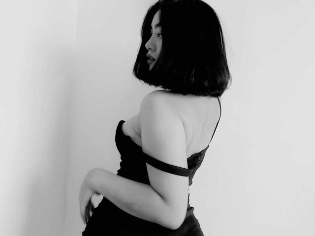 Black-and-white photo of Annakomatsu with shoulder-length dark hair, smiling, wearing a strapless black dress, exposing her tits, with one arm resting on her ass.