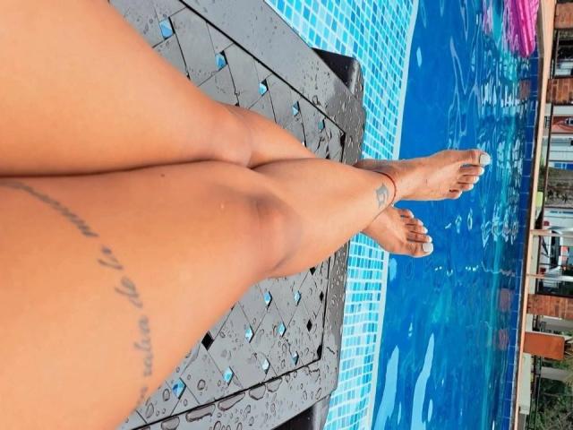 FiorellaBossh's legs with a tattoo on the left thigh, feet in a blue-tiled pool, toes painted white, on a black metal poolside, water droplets on her skin.