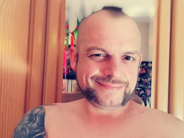 DonGranato69, bald with a shaved head and beard, smiling with a nose ring, tattoo on left shoulder, shirtless, in front of wooden door, looking slightly to the right.