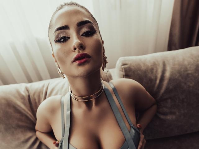 LoraDevine with blonde updo, heavy eyeliner, red lipstick, gold earrings, choker, and blue top, hands on her tits, sitting on beige couch, soft light from window.