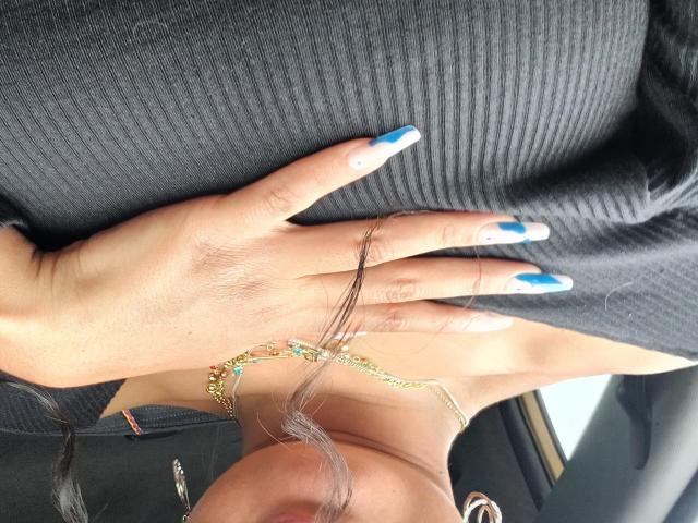 ArielleWilde's hand with blue and white nail polish rests on her ribbed black top, showing a gold chain bracelet with small blue gems. Her face is partially visible.
