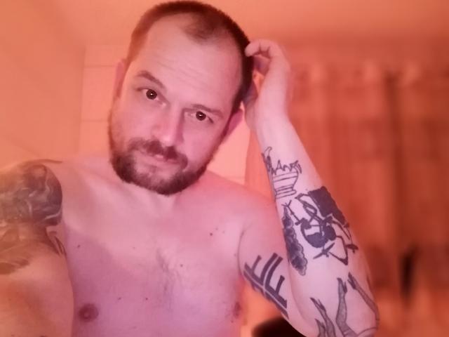 Shirtless DonGranato69 with tattoos, bald with a beard, holding his head, looking at the camera, in a warm orange-lit bathroom.