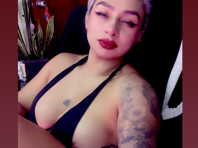 AvrilGray with short purple hair, wearing a black halter top, exposing her big tits, has a tattoo on her chest, a flower tattoo on her right arm, and red lipstick.