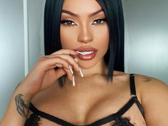 JunneLiu with black straight hair, brown eyes, full lips, wearing black lace bra, hand near mouth with white nail, tattoo on right shoulder, medium tits, smiling.