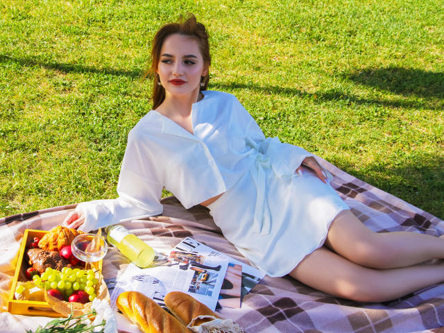 CamillaRoberts is lying on a plaid blanket with her ass up, wearing a white dress, surrounded by a picnic with grapes, bread, and a yellow drink.