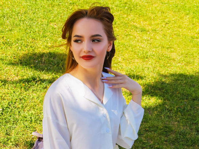 CamillaRoberts with red lipstick, white button-up shirt, and green grass background, has her hand near her neck, brown hair in a half-updo, and a slight smile.