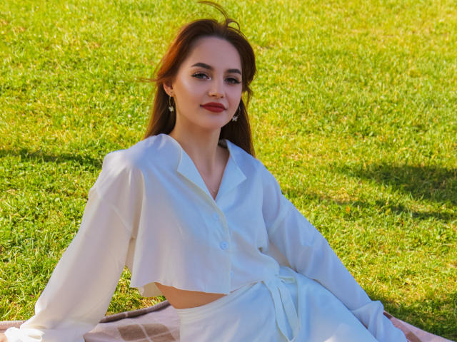CamillaRoberts with red hair, white blouse, and white skirt, sitting on grass, legs crossed, smiling, showing her tits, with earrings, and a pink checkered blanket.