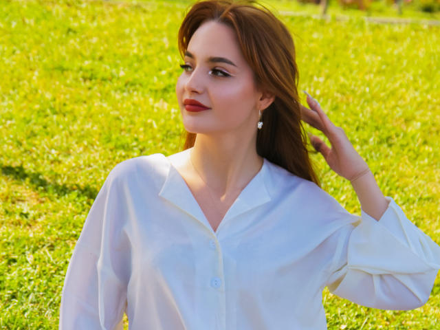 CamillaRoberts with long brown hair, wearing a white button-up shirt, has a hand in her hair, smiling with red lipstick, small white earrings, green grass background, sunlight.