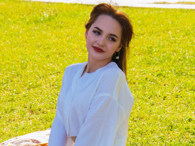 CamillaRoberts with red lipstick, white blouse, and green grass, smiling slightly, looking to the side, brown hair up, small earrings, sitting on a blanket, sunlight on grass.