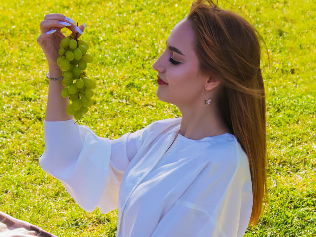 CamillaRoberts with red hair, white dress, holding green grapes, side profile, smiling, earrings, sunny grass, right arm raised, soft light, relaxed, pretty face, gentle expression.