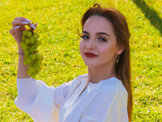 CamillaRoberts with red hair holding green grapes, wearing a white shirt, smiling, red lipstick, earrings, sunlight, grass. Her tits are subtle under the shirt.