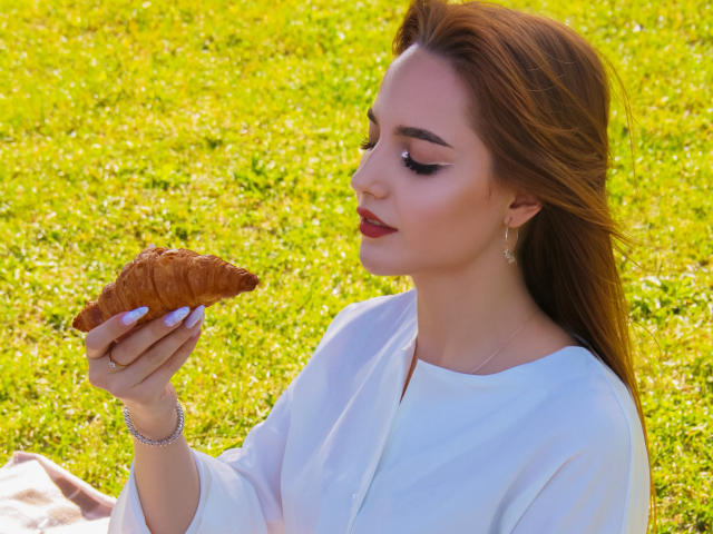 CamillaRoberts with red hair, white shirt, holding a croissant, green grass background, wearing earrings, red lipstick, purple nails, bracelet, looking down at the croissant, sunny day.